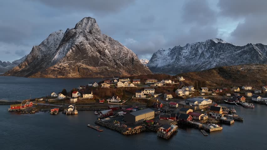 Aerial view of Reine's colorful houses nestling by the fjord, contrasted against the snow-dusted mountains under a dramatic sky, Reine, Norway.