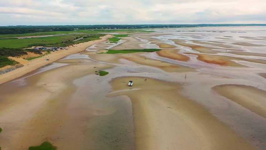 Aerial view of expansive sand flats at low tide where a stranded boat sits among tide pools, sculpted ridges, and patches of green marsh grass stretching across the coastal landscape.