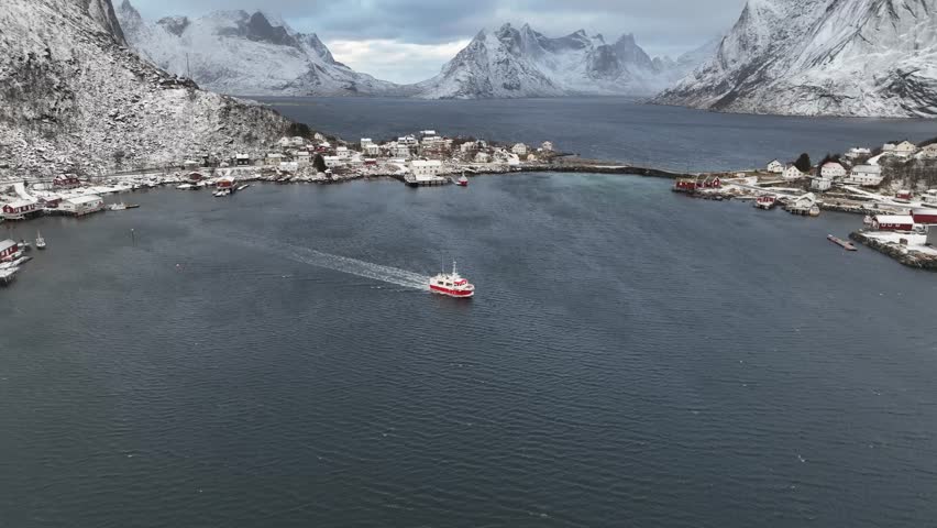 Aerial view of a red and white boat sailing on the dark blue waters near Reine village under snow-capped mountains and cloudy sky, Reine, Norway.