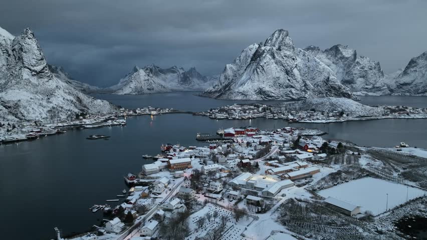 Aerial view of snow-covered Reine village nestling by the bay, crowned by jagged mountains under a dramatic sky, showcasing the stark beauty, Reine, Norway.
