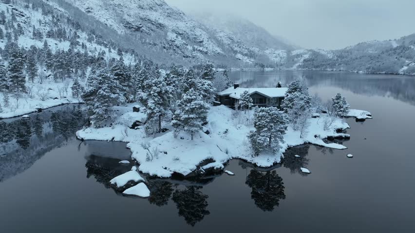 Aerial view of snow-covered houses and trees near a tranquil lake, reflecting the serene landscape in its dark waters, Norway.