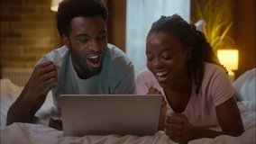 Overjoyed happy African American family ethnic couple woman man lying bed watching digital tablet online sport match betting giving high five gesture support victory winning luck success home bedroom - Powered by Shutterstock - Get 15% off with code: PIKWIZARD15