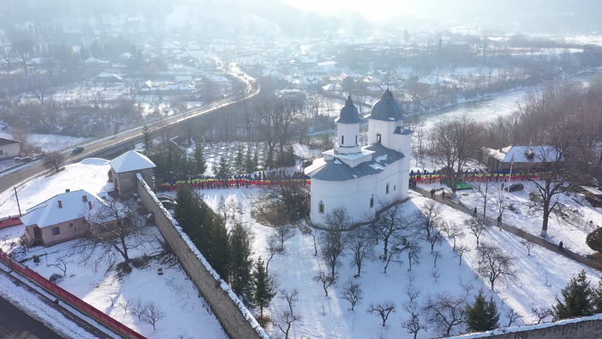 An ascending aerial shot slowly reveals Raducanu Monastery in Targu Ocna, Romania. A long line of people holds a massive Romanian flag on the snow-covered grounds during a winter national event.
