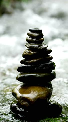 A vertical close up video shows a hand carefully placing a small stone on top of a balanced rock stack. The cairn sits in a flowing river.