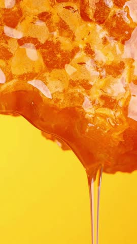 A close up vertical video shows thick golden honey dripping slowly from a natural honeycomb. The vibrant yellow background emphasizes the pure, sweet liquid.