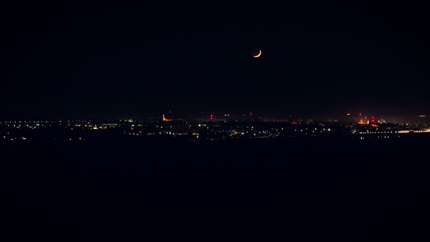 Crescent moon shining above city skyline at night. Lunar glow rising over sparkling urban lights. Thin moon hovering above buildings in deep darkness. Evening city glowing softly under moonlit sky