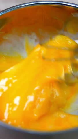 A close up vertical video shows a wire whisk rapidly mixing vibrant yellow egg yolks in a metal bowl, preparing ingredients for a recipe.