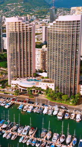 Aerial view of Honolulu, Oahu. Marina with sailboats, two tall towers, rooftop pool, and lush mountain backdrop. Vertical video.