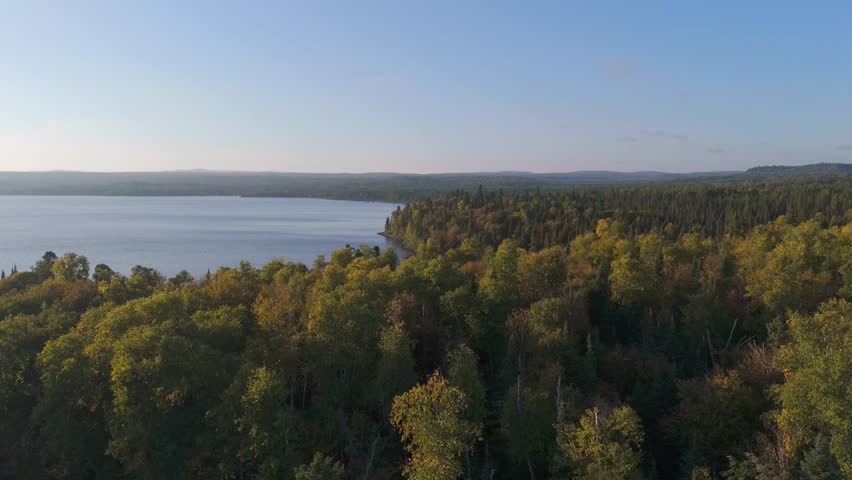 A warm golden hour scene over the Ontario side of Lake Superior, where sunlight bathes the forest in rich, glowing tones and highlights the natural beauty of the shoreline. Aerial view