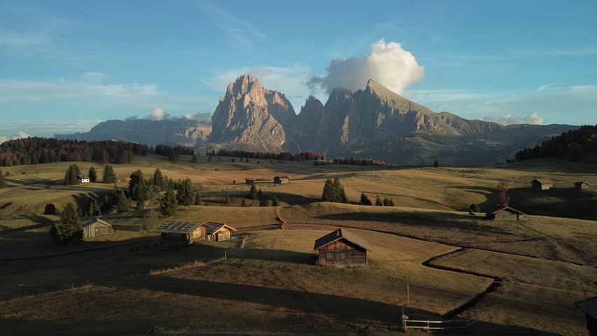 Aerial view of golden fields meeting rugged mountains and quaint wooden huts, creating a serene landscape panorama, Seiser Alm, Trentino-Alto Adige, Italy.