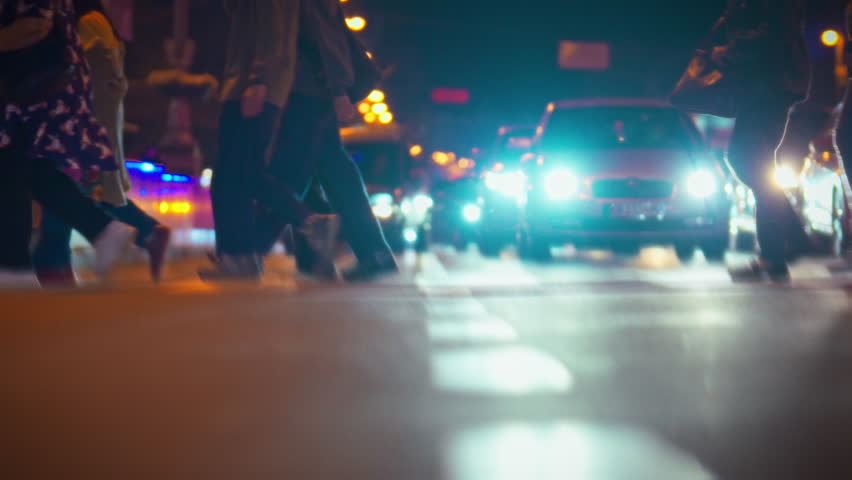 Crowds of pedestrians are crossing a lively street under the glow of streetlights at night. Vehicles navigate around them while city life buzzes with energy and movement
