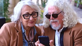 Happy senior couple sitting together at outdoor cafe using smartphone and looking at social media. Elderly people enjoying technology, connection, communication, digital lifestyle, love and leisure - Powered by Shutterstock - Get 15% off with code: PIKWIZARD15