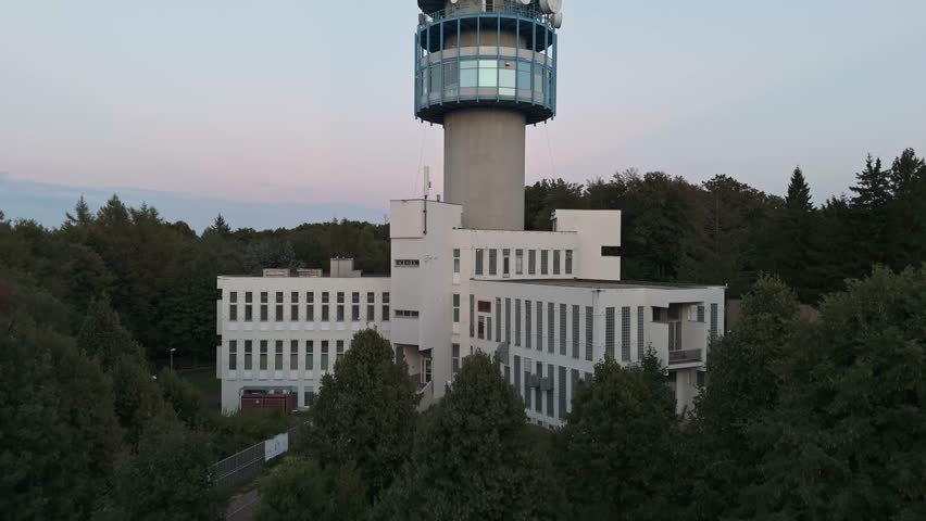 Ascending aerial view along the Kékes TV Tower from the base building towards the top, featuring various antennas with the hazy Mátra Mountains in the background in Hungary.