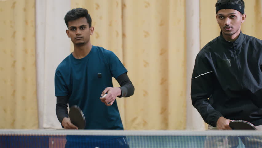 Two siblings engaged in competitive table tennis rally, one in black top striking ball into net while brother in blue focuses on return, showcasing energy and coordination