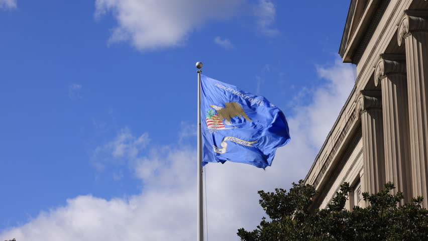 The Department of Justice flag, featuring a golden eagle, waves dynamically in the wind against a bright blue sky with clouds. A portion of the RFK Building is visible.