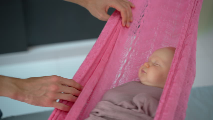 Loving father gently swings his swaddled two-month-old baby in a hammock to help him fall asleep, creating a peaceful and nurturing family bedtime moment.