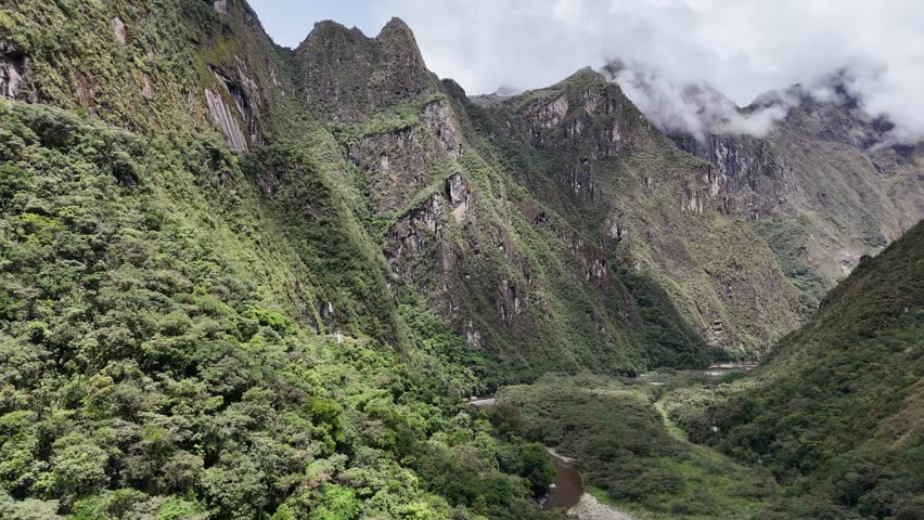 A majestic shot rises vertically from the lush banks of the Urubamba River near Aguas Calientes, dramatically revealing the numerous iconic mountain peaks of the Sacred Valley