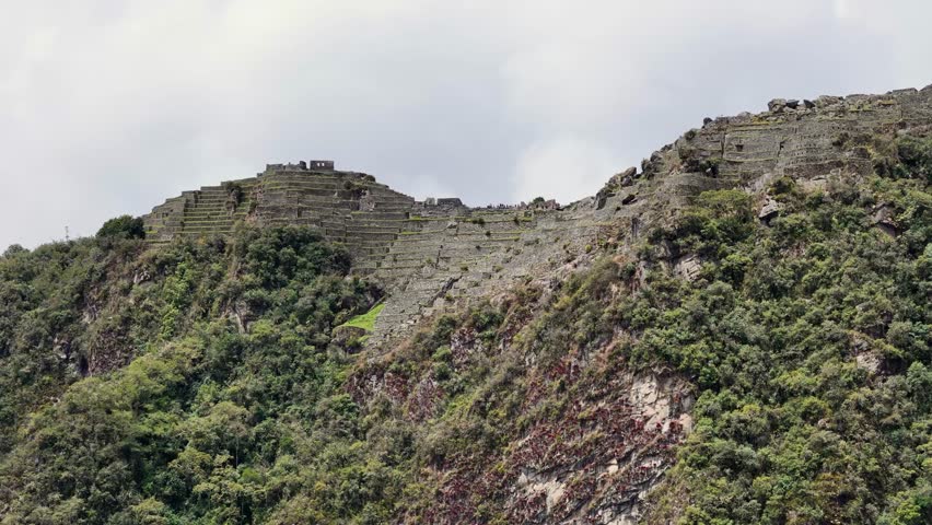 From mountainside circles directly opposite urban area of Machu Picchu, capturing the ancient structures and terraces with dramatic clouds drifting and swirling behind the upper levels of the citadel