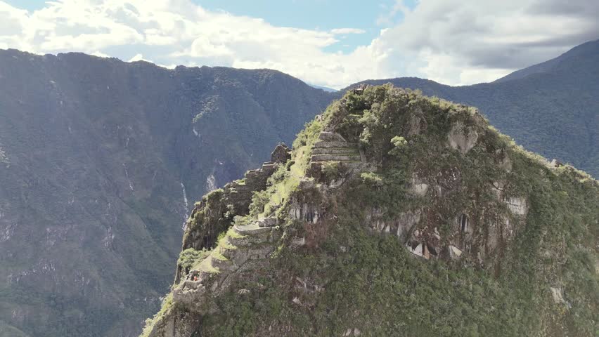 A dynamic aerial shot circles closely around the steep peak of Huayna Picchu, revealing the ancient stone structures built on its different levels and the narrow stairs leading to the summit