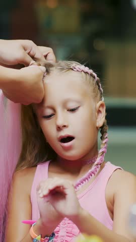 Beautiful blond girl, of seven years old, braided two pigtails, do a hairstyle with pink locks of hair in a beauty salon, a hairdresser