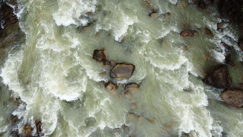 Mountain river with fast flowing, rapids and narrow wooden footbridge in overcast rainy day, aerial vertical view down while moving above the river downstream
