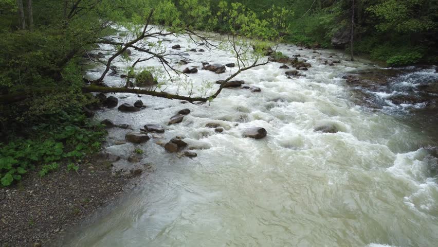 Mountain river with fast flowing, rapids, tributary stream and narrow wooden footbridge in overcast rainy day, aerial view while moving backwards above the river downstream
