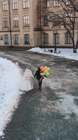 winter wedding. Aerial view, newlywed couple in wedding dresses are runing with colorful balloons through the snow-covered park, against the background of ancient architecture and paving stones