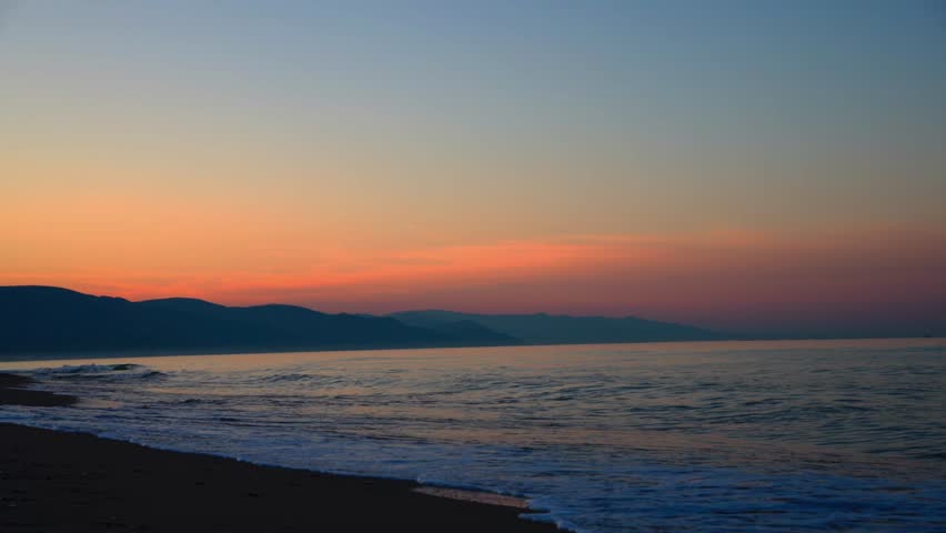 Coastal sunset scene with tranquil sea waves, dark beach sand and layered mountains on horizon. Beautiful golden hour seascape and landscape