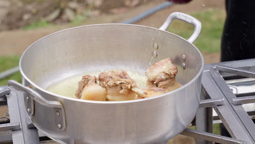 Pork pieces frying in hot oil inside a large aluminum pot. A chef covers the pot with a red lid while cooking outdoors at a country restaurant