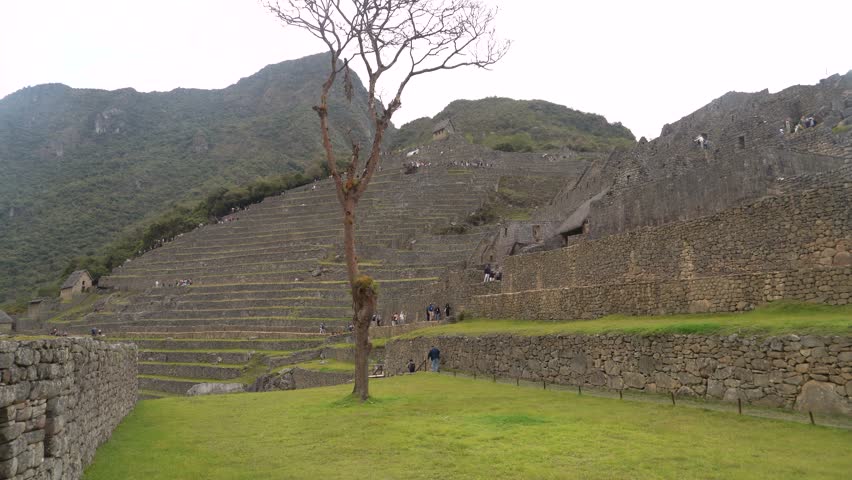 Western tourists walking through the agricultural and urban sectors of Machu Picchu, specifically near the iconic Temple of the Sun and Royal Tomb area, surrounded by ancient stone walls, rooms