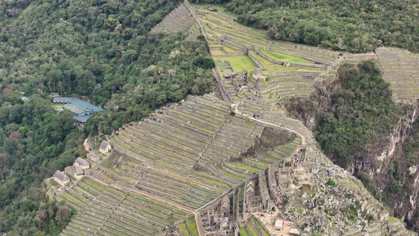 A close-up aerial shot moves from upper terraces of Machu Picchu down to the lower levels, with tourists visible roaming the site, before continues to pull back, flying past the iconic Huayna Picchu