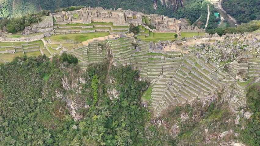 A complex and cinematic drone shot looks down on the terraces of Machu Picchu, slowly circling the site while simultaneously pulling back to reveal more ancient stone structures, the iconic peak, and 