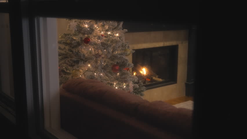 Fireplace burning next to a Christmas tree at home, while snow falling outside. Dolly shot, slow motion.