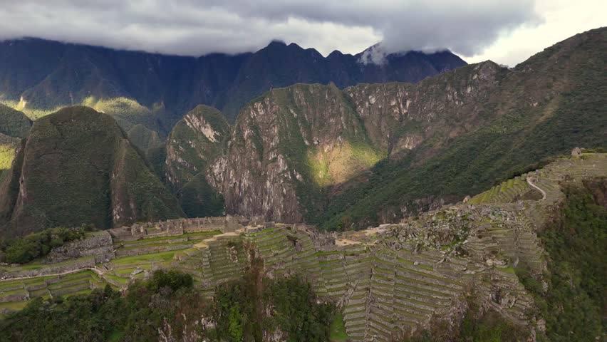 A smooth drone shot pulls away and flies downward from the green terraces of Machu Picchu, dramatically revealing the immense scale of the ancient citadel and its surrounding mountain landscape