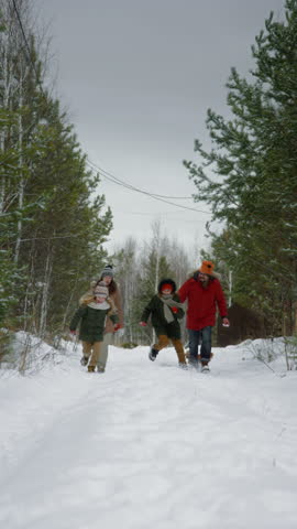 Full vertical shot of young Caucasian family with pet maltipoo dog running and throwing snowballs in forest. Father with missing limb disability playing in snow with wife and kids