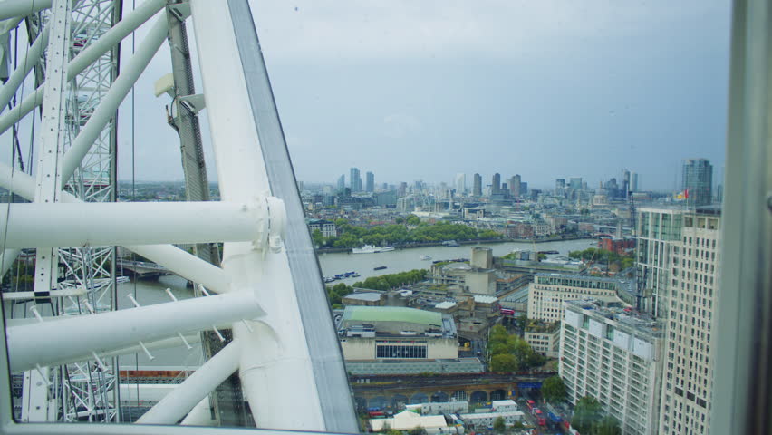 Aerial view of London seen through the metal frame of a large observation wheel, showing the River Thames, city skyline, and boats under a cloudy, rain-speckled sky.