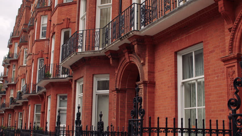 Row of traditional red brick London townhouses with ornate iron railings, arched entrances, and black wrought-iron balconies under a cloudy sky.