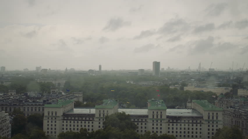 Aerial view of London under overcast skies, featuring government buildings with green roofs, dense trees, cranes, and high-rises in the misty, rain-soaked distance.