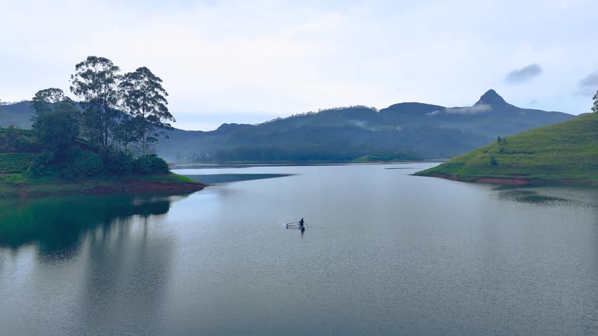 Aerial view of Maussakelle Reservoir with Sri Pada (Adam’s Peak) in the background, Sri Lanka. Tranquil drone landscape of calm lake, misty mountains, and a lone fisherman at dawn.