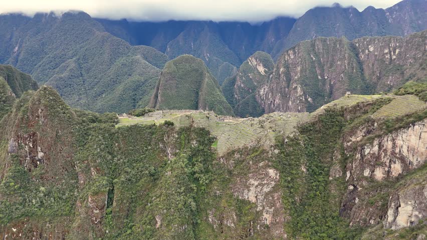 A smooth aerial shot flies forward towards the Machu Picchu citadel, gradually revealing its green terraces, ancient buildings, and precise location perched on a cliff amidst the surrounding mountain