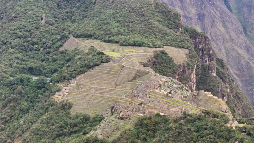 Aerial shot moves forward towards iconic Intihuatana stone and surrounding terraces. Camera pans down close-up view, revealing tourists wandering among Nusta