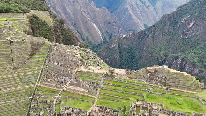 A stunning shot begins with a close-up on the iconic green terraces and stone structures of Machu Picchu, pulls back and soars upward to reveal the entire ancient citadel, surrounded by majestic cloud