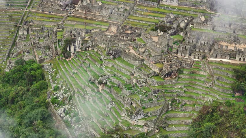 A dynamic drone shot flies in close over Machu Picchu, then rises vertically through misty clouds to reveal the different levels and layers of the ancient stone structures built into the mountainside