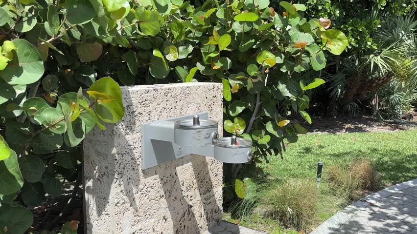 A stone water fountain with metal fixtures is installed beside a paved path surrounded by green plants and grass in Palm Beach, Florida.