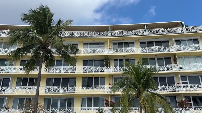 A multi-story yellow apartment building with balconies and palm trees in front is seen under a partly cloudy sky in Palm Beach, Florida.