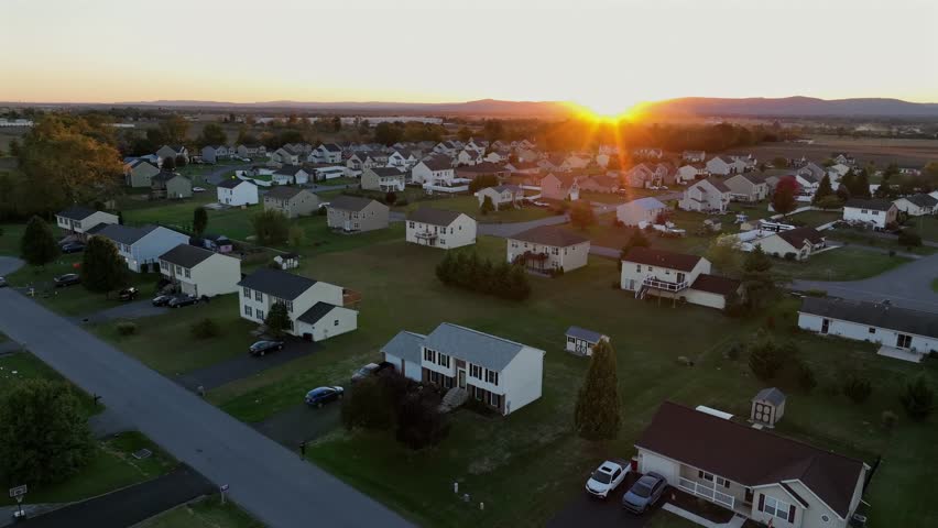 Single family two-story homes in quiet suburb neighborhood of american town. Golden sunset behind hills of Pennsylvania in distance. Aerial raising wide shot. Late summer days in USA.