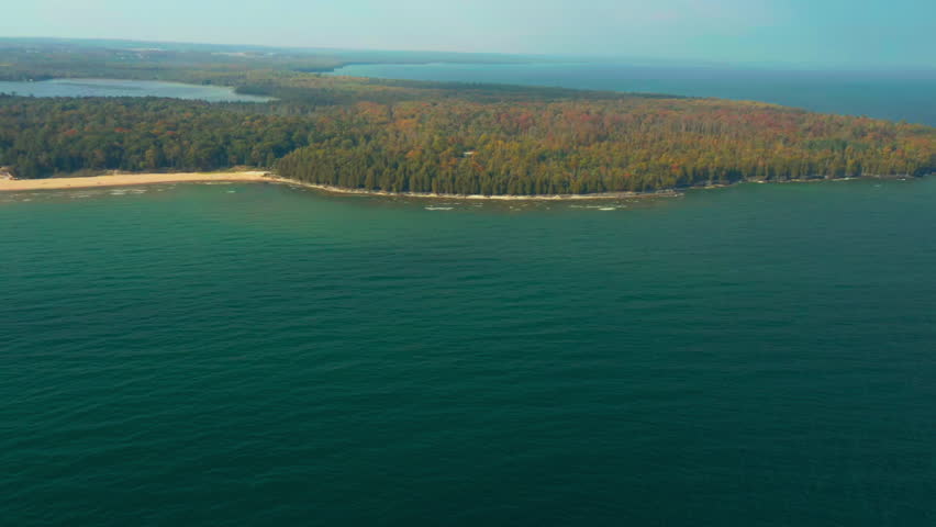 The scene shows the meeting of deep green forest and the clear, blue-green waters of Lake Michigan, where sandy beaches curve gently along the edge of the peninsula in early autumn.