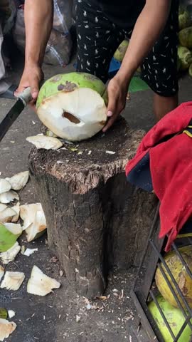 Man chopping coconut with machete on wood stump in outdoor market setting