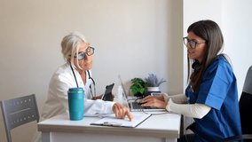 Senior female physician in a white coat guiding a smiling young nurse in blue scrubs, both pointing at a clipboard and working together on a laptop in a modern medical clinic office - Powered by Shutterstock - Get 15% off with code: PIKWIZARD15