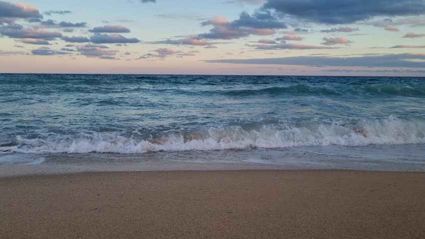 Powerful sea waves crash on the sandy beach at sunset, with colorful clouds and deep blue water creating a dramatic and peaceful ocean scene.
